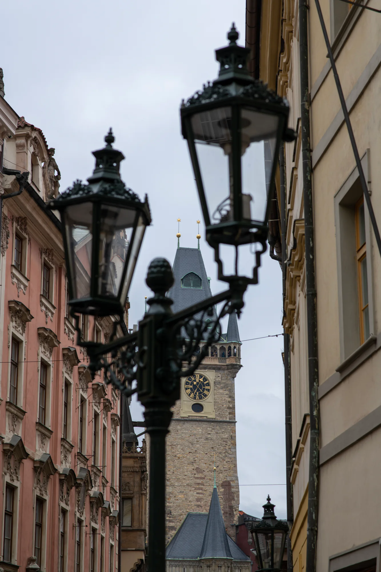 Old Town Hall tower and astronomical clock seen between historic buildings and ornate street lamps in Prague.
