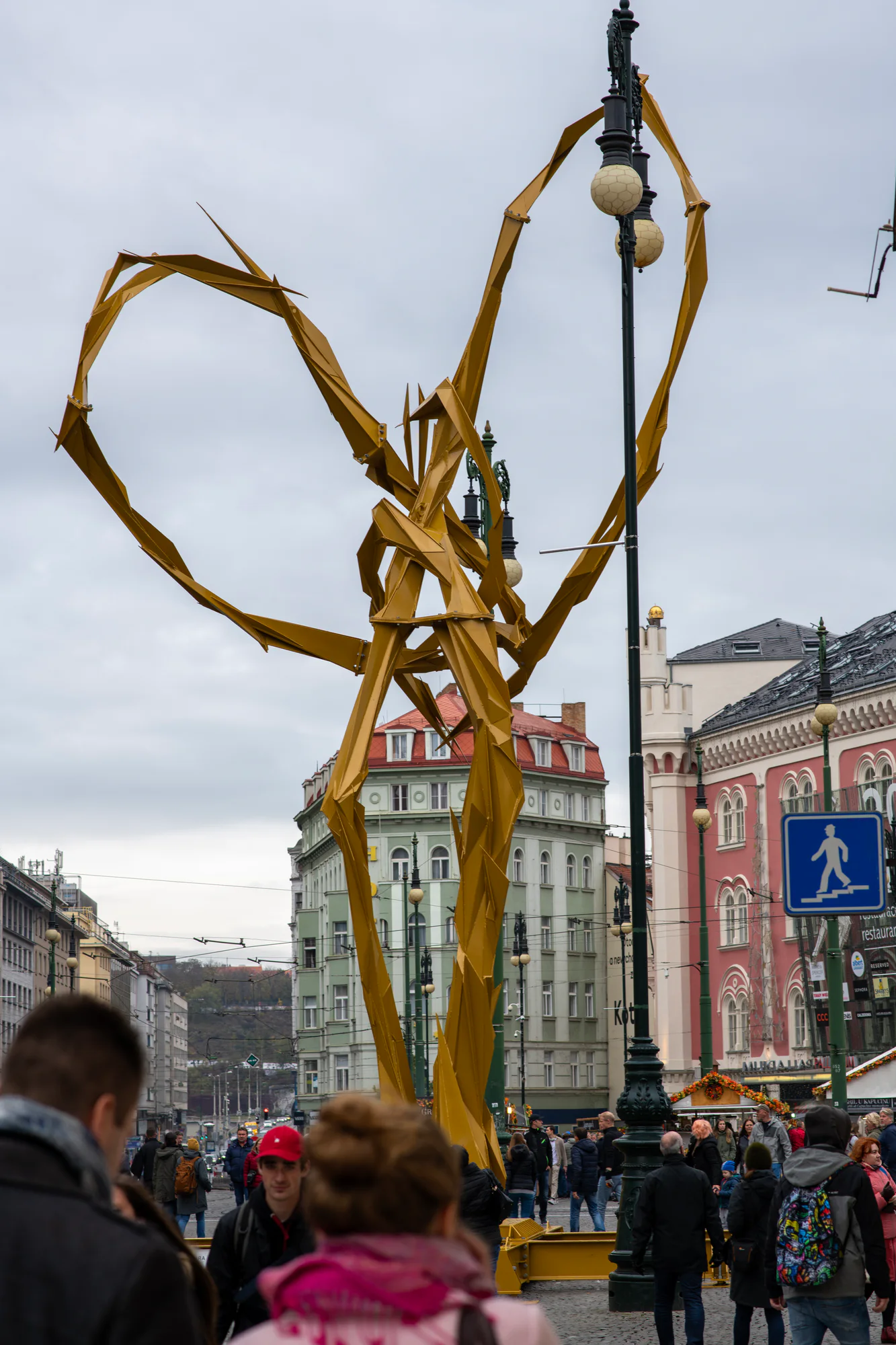 Monumental golden geometric sculpture rising above pedestrians on a street in central Prague.